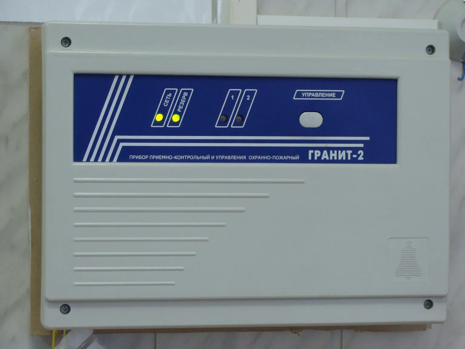 Прибор «Гранит-2» на стене: на синей панели надпись «прибор приёмно-контрольный и управления охранно-пожарный», горят лампы «Сеть» и «Резерв», лампы шлейфов 1 и 2 погашены, справа кнопка «Управление». Фото: Georg Pik, общественное достояние